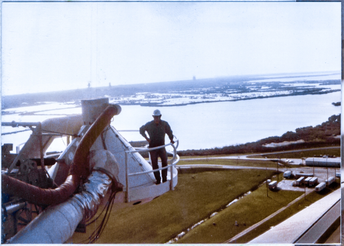 Cantilevered 270 feet above the land and water behind him, James MacLaren poses on the Gaseous Oxygen Vent Hood at Space Shuttle Launch Complex 39-A, Kennedy Space Center, Florida. In the far distance, in silhouette, structures of the Titan III, Integrate-Transfer-Launch area on Cape Canaveral Air Force Station can be seen. From left to right: Launch Complex 41 Mobile Service Tower and Umbilical Tower, Launch Complex 40. Pad 41 is where the two Voyager Spacecraft were launched from, on their endless journey into interstellar space, and is also where the two Viking Spacecraft were launched from, on their journey to become the first objects to successfully land on the planet Mars.